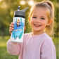 A young girl with blonde hair in a ponytail smiles while holding a custom white water bottle outdoors. The water bottle features a printed illustration of a gymnast on a swing with the name 'Lexi' written below in black script.