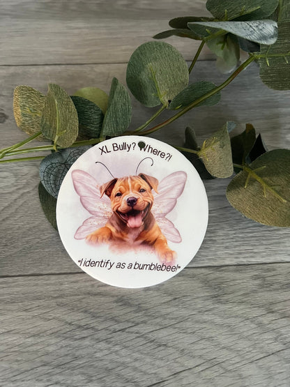 Round sticker with a dog wearing fairy wings on a wooden surface with green leaves.