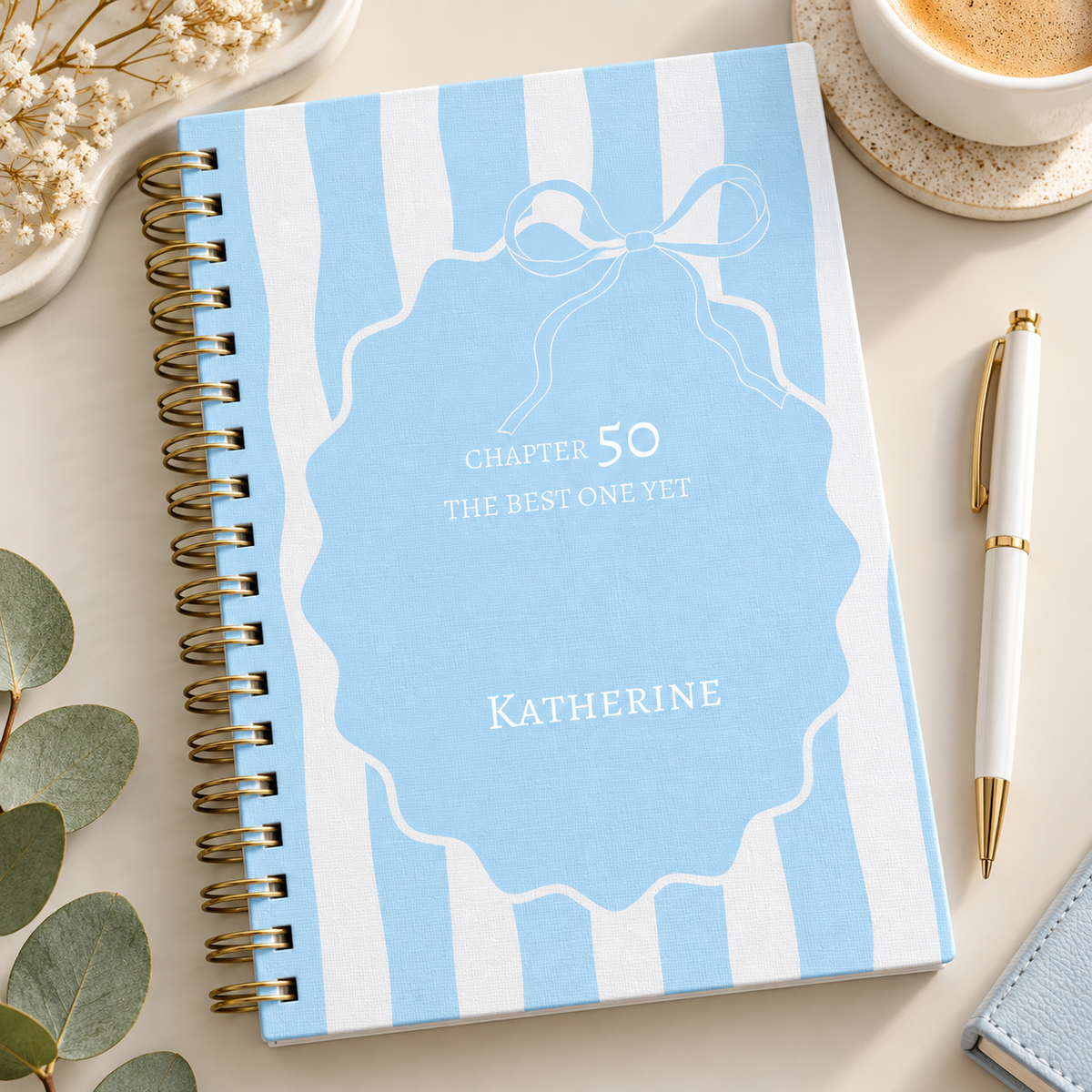 A spiral-bound notebook with a light blue and white striped cover, featuring a central blue label design with the text 'Chapter 50 The Best One Yet' and the name 'Katherine.' It is styled on a light beige surface with a white pen, a cup of coffee, dried flowers, and green leaves around it.