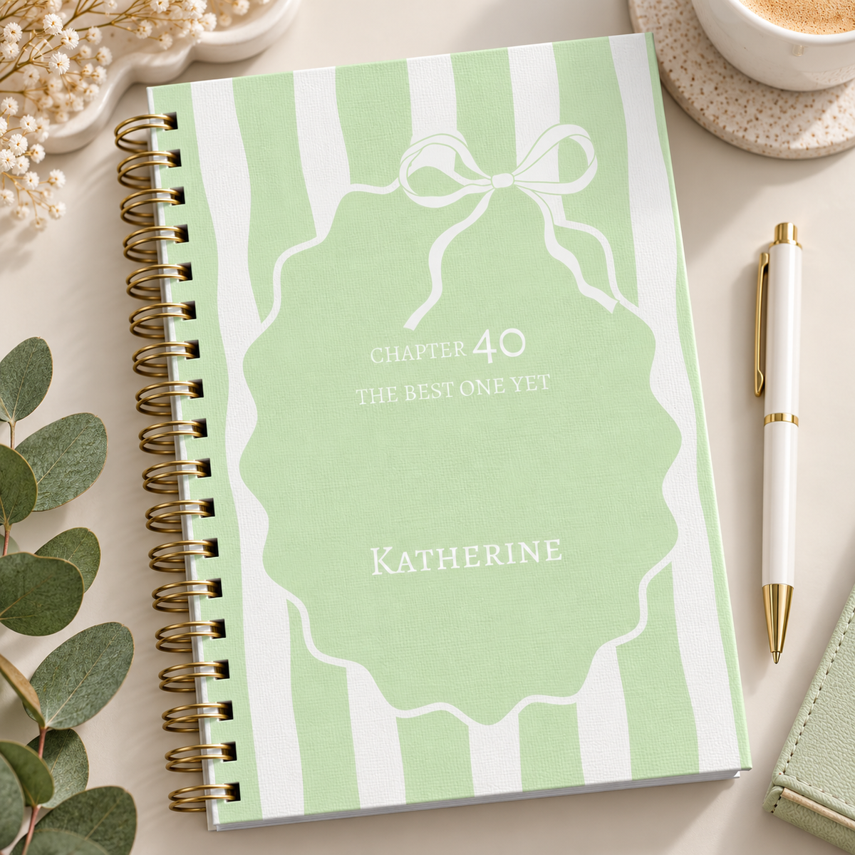 A personalized spiral-bound journal with a mint green and white striped cover, featuring a white outline of an ornament with the text 'Chapter 40 The Best One Yet' and the name 'Katherine'. The journal is styled in a flat-lay arrangement with a gold pen, a cup of coffee, eucalyptus leaves, and baby's breath flowers on a light beige surface.