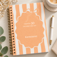 A spiral-bound notebook with a peach and white striped cover, featuring a central peach label with a bow outline and the text 'CHAPTER 30 THE BEST ONE YET' and the name 'KATHERINE'. The notebook is styled on a light surface with a pen, a cup of coffee, flowers, and green leaves.