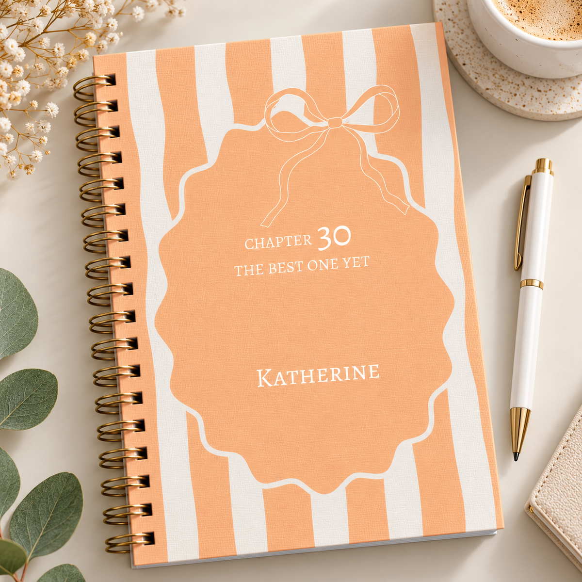 A spiral-bound notebook with a peach and white striped cover, featuring a central peach label with a bow outline and the text 'CHAPTER 30 THE BEST ONE YET' and the name 'KATHERINE'. The notebook is styled on a light surface with a pen, a cup of coffee, flowers, and green leaves.