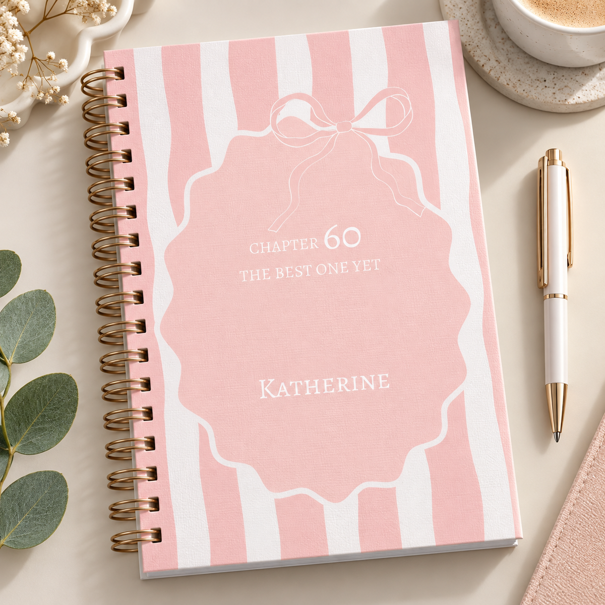 A spiral-bound notebook with a pink and white striped cover, featuring a central pink badge design with the text 'CHAPTER 60 THE BEST ONE YET' and the name 'KATHERINE'. The notebook is styled on a beige surface with a gold pen, a cup of coffee, eucalyptus leaves, and dried flowers.