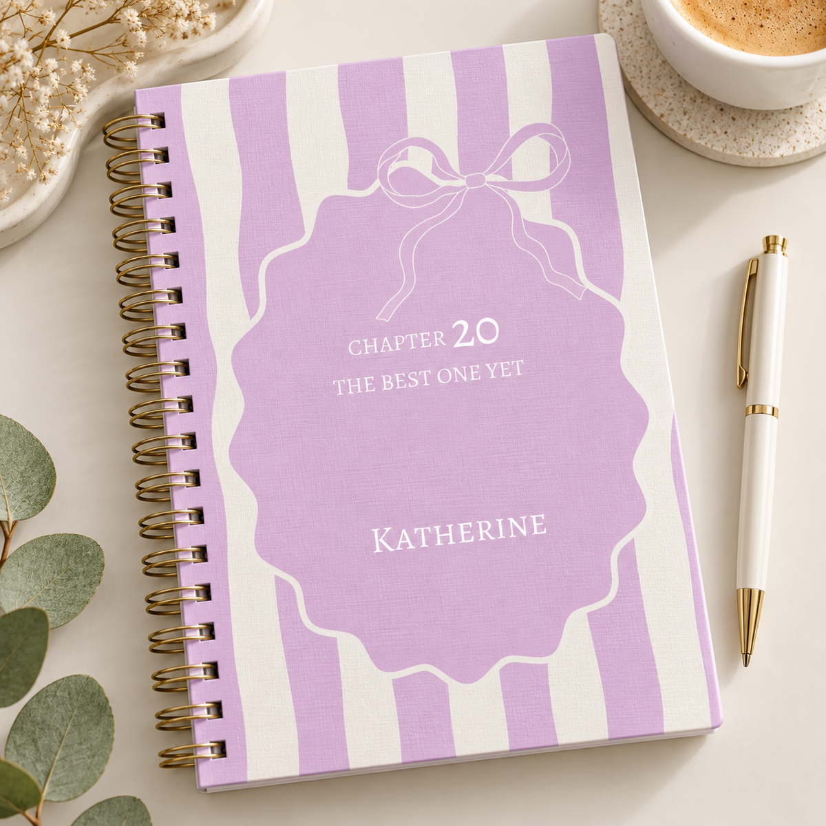 A spiral-bound notebook with a lavender and white striped cover, personalized with the text 'CHAPTER 20 THE BEST ONE YET' and the name 'KATHERINE' on the front cover. The notebook is styled on a light beige surface with a gold pen, a cup of coffee, dried flowers, and green leaves.
